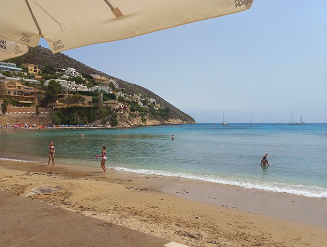 Going to El Portet Beach, one of the best things to do in Moraira several people on a sandy beach with crystal clear water and villas on a hill in the background