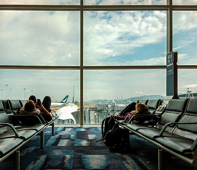 2 people lying on benches at an airport trying to sleep, with a plane behind the window