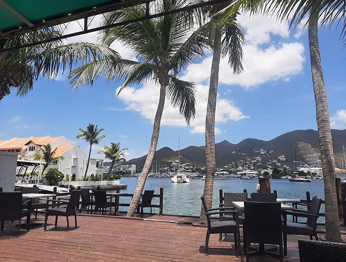 Living in St Maartem chairs, tables and palm trees on a terrace overlooking a bay with bright blue water and several boats, with a blue sky and green mountains in the background