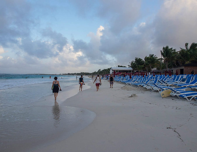 A long sandy beach with several people and several rows of sun loungers plus palm trees in the background