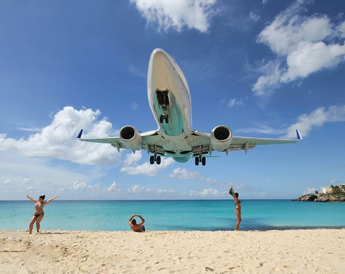 A large plain flying very low over a sandy beach with 3 people on the beach