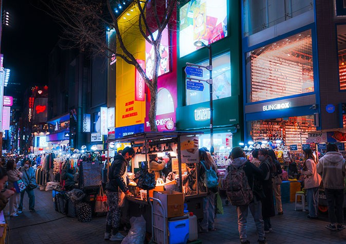 Popular neighborhoods to experience in Seoul A busy shopping street at night with brightly lit stores, billboards and street food