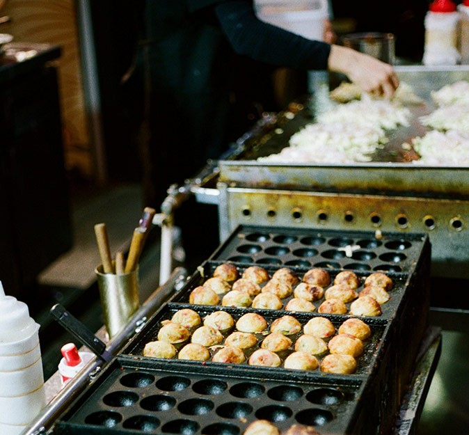 A foodstall with small brown balls being cooked on a black metal grill
