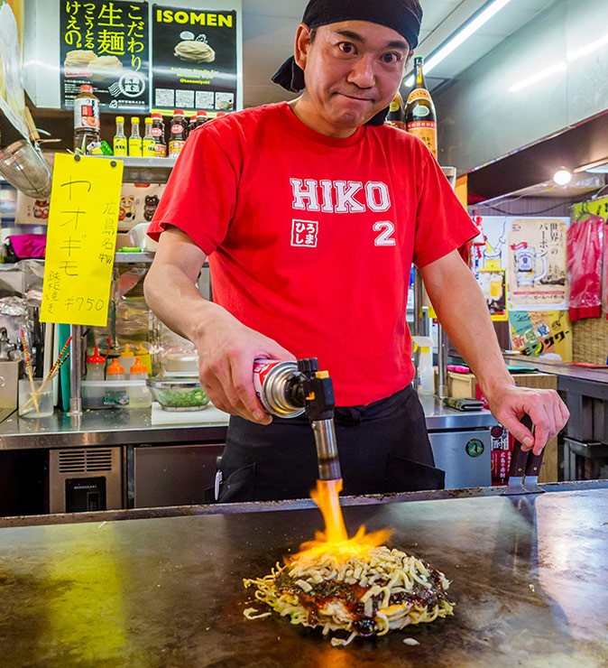 A man in a red t-shirt holding a small flamethrower above a pancake on a grill