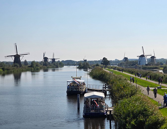 One of the best places to visit in the Netherlands A canal with 2 boats, and several windmills and several people on both shores