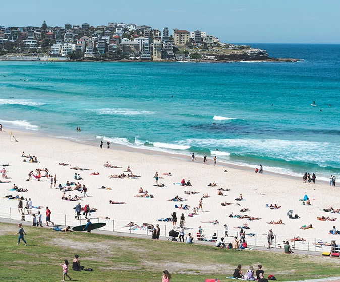Things to know before going to the beach in Sydney A white sandy beach with several people, blue ocean and buildings in the background