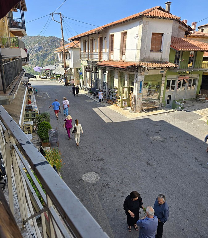 A greek town seen from a top floor balcony, showing people walking in a small street with buildings with orange roofs, several housing small shops and cafes