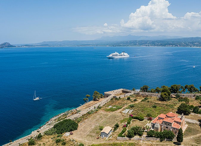 Why you should take a cruise Green and brown coastline with a sailboat and a cruise ship in the background, surrounded by blue water
