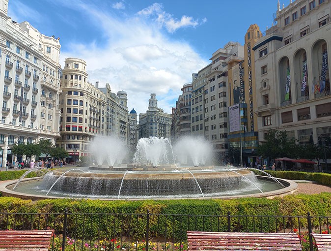 A large fountain with benches in front of it and historic buildings in the background