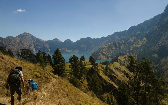 2 people hiking on a steep cliff with mountains and a lake in the background