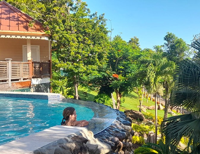 What should a hotel room have? I love a hotel with a pool! A woman leaning on the edge of an infinity pool with part of a hotel room in the background, surrounded by palm trees and greenery