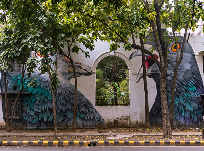 Street art made up of 2 large pigeons on a white wall with several trees in front of it