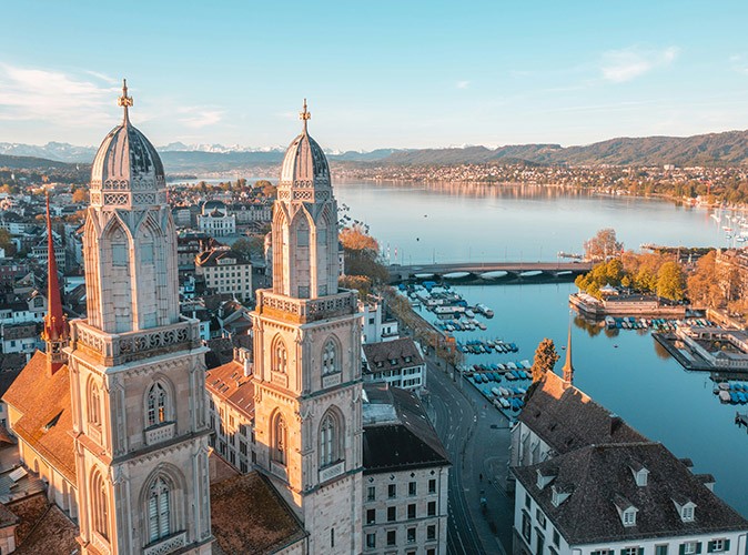 How is life in Zurich An aerial view of Zurich showing a historic church, several houses, a lake with small boats and mountains in the background