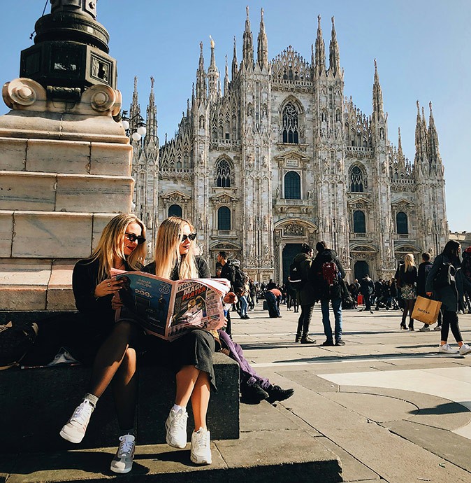2 blond girls sitting on a stone bench holding a news paper, with a large cathedral in the background