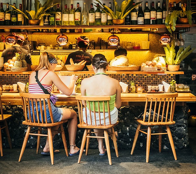 Digital nomads in Ubud, Bali 2 girls sitting on wooden chairs inside a restaurant with warm yellow lighting and several bottles plus plants on display against a wall