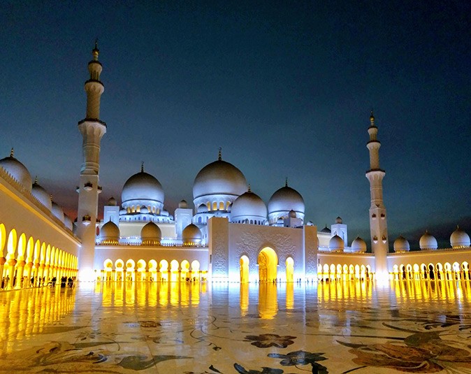 One of the best day trips from Dubai: visit Abu Dhabi A large white mosque at night with warm yellos lighting and a large marble floor in front of it