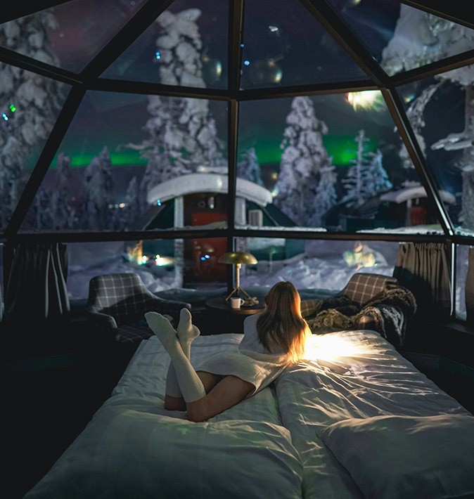 A blond woman lying on a white bed in a Finnish transparent igloo with a view of snowy trees and the northern lights