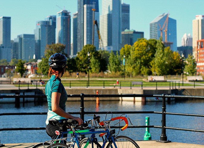 Things to know before traveling to Canada A woman holding 2 bicycles looking out over a river with a city with modern skyscrapers in the background