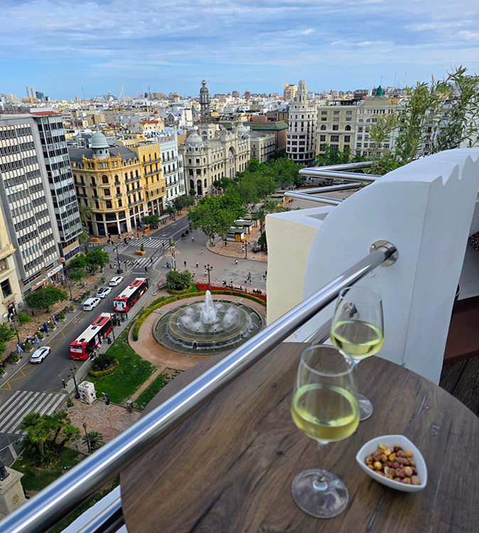A city center rooftop bar in Valencia 2 white wine glasses and some peanuts on a brown table ona rooftop overlooking a city square with a fountain, historical buildings and buses