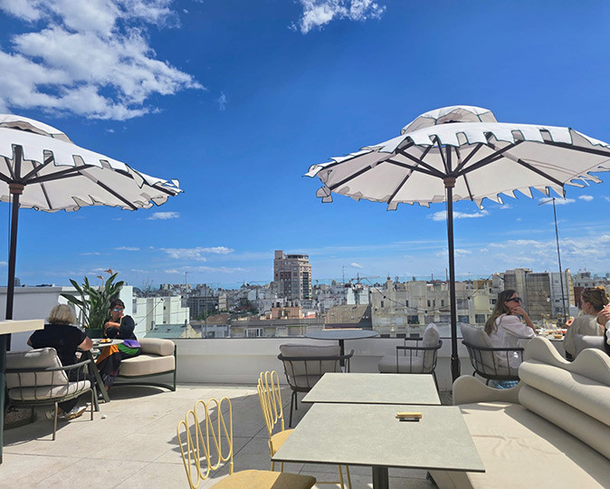 A rooftop bar overlooking the Valencia city skyline white tables, chairs and 2 umbrellas on a rooftop with several people and a city skyline