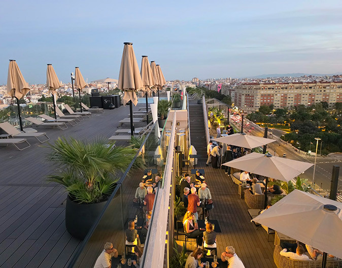 Skybar Valencia at Novotel A rooftop bar in Valencia showing stylish tables with people and an area with sun loungers and umbrellas, surrouned by the skyline of Valencia
