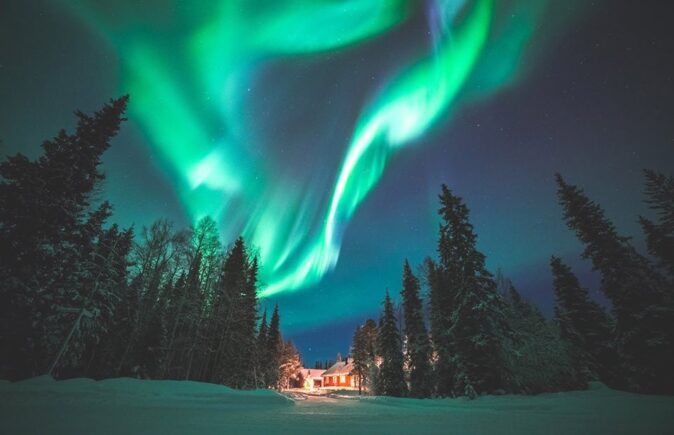 a once in a lifetime trip to see the northern lights northern lights above pine trees surrounded by snow with a cottage in the background