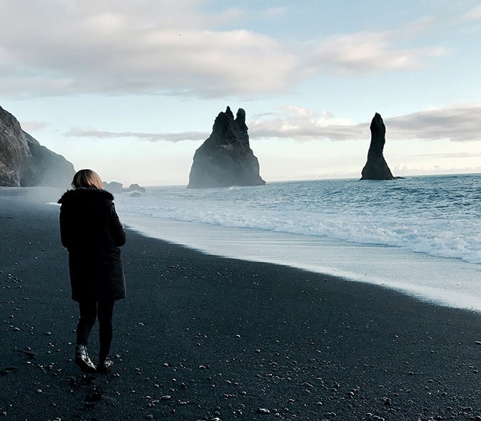 A woman in a thick black coat standing on a black sand beach with rock formations in the background