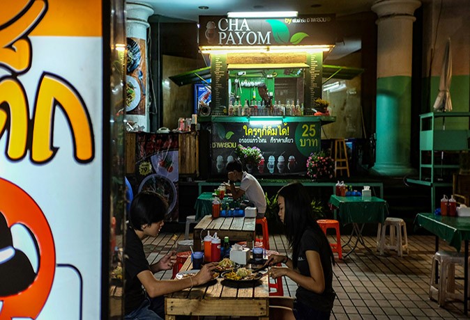 A dimly lit, simple, Thai restaurant in Bangkok with a few people sitting at small tables with small plastic chairs eating Thai food