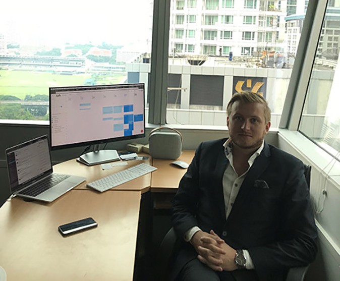 A man in a black suit sitting behind a desk with a computer screen, with large windows in the background