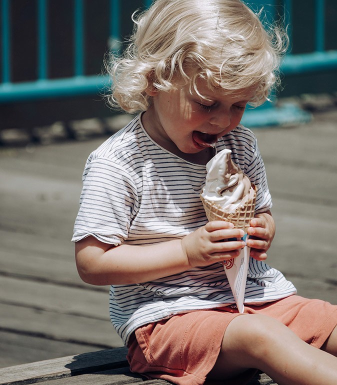 A blond child sitting on a wooden floor eating a large icecream