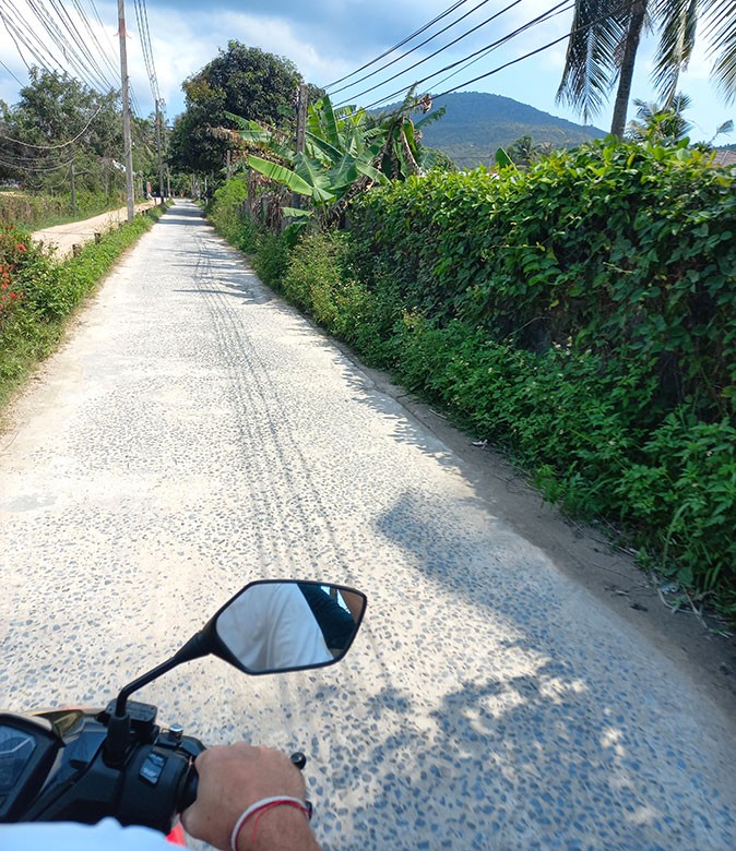 Tips for safely visiting Thailand A long, white sandy road with greenery and palm trees on the side, with part of a scooter and a hand showing in one corner