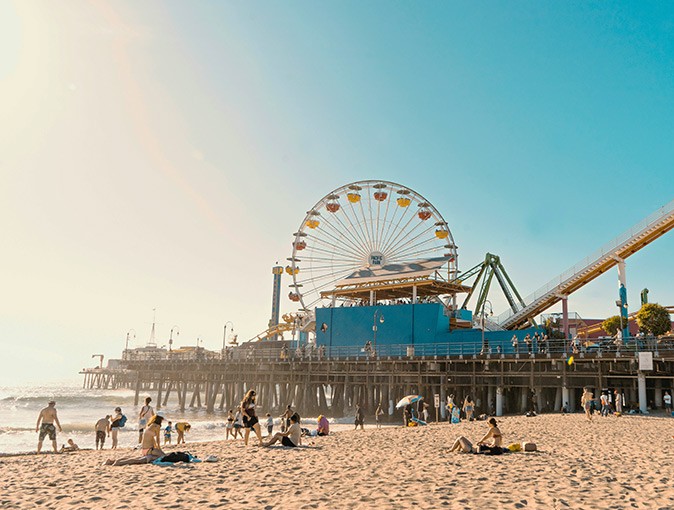 A sandy beach with several people with a large pier with a ferris wheel in the background