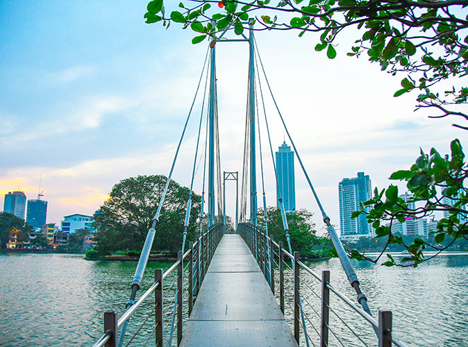 Life in Colombo A suspension bridge over a body of water with some trees and highrise buildings in the background