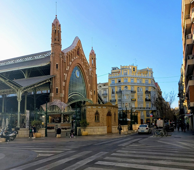 A historic Spanish market hall with roads in front of it, a car, a few pedestrians and apartment buildings