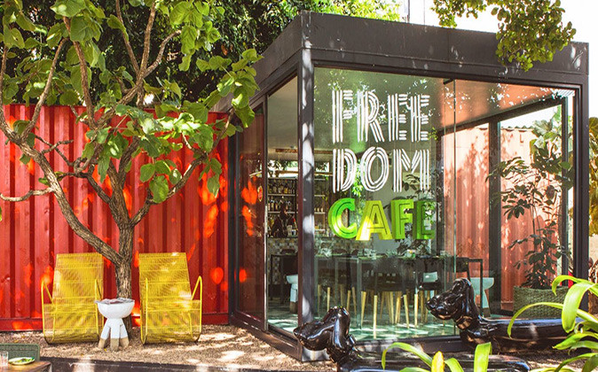 A cafe exterior showing part of a red shipping container and a modern, glass entrance plus bright yellow chairs