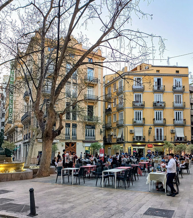 Valencia as a place for retirement A city square in Valencia with a restaurant terrace, various people, a tree and 2 historic apartment buildings