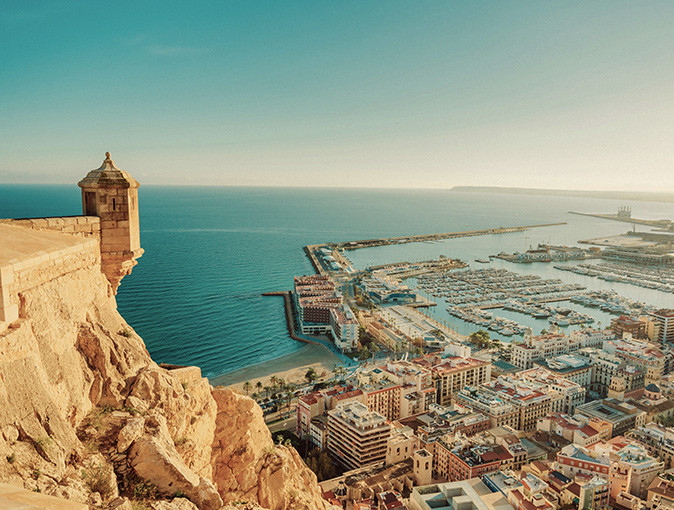 Aerial view of Alicante showing part of the castle overlooking city buildings and a large marina with the sea in the background