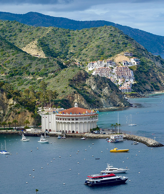 Planning a day trip to Catalina Island, California Aerial view of Catalina Island showing a ruggid coastline with several boats, a breakwater with a large white building and green hills with some houses in the background