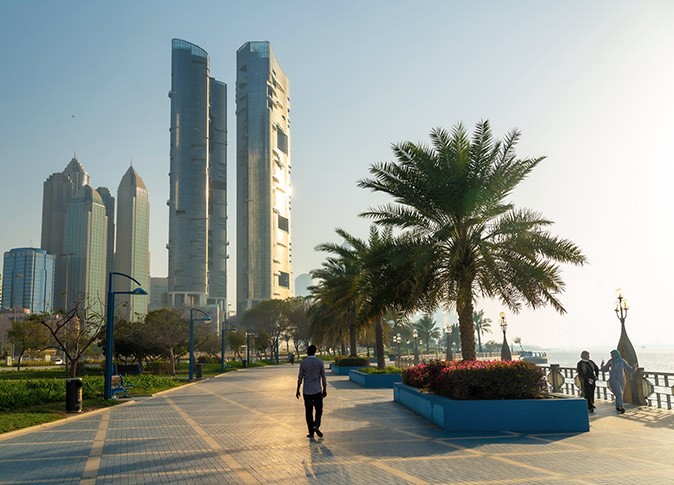 A wide, paved boardwalk with palm trees, 3 people walking and high rise buildings in the background