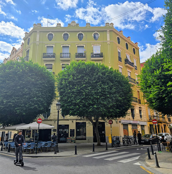 one of the best neighborhoods to stay in Valencia: Ruzafa colorful historic buildings with several trees on the sidewalk, a terrace and someone on an electric scooter passing by on the road