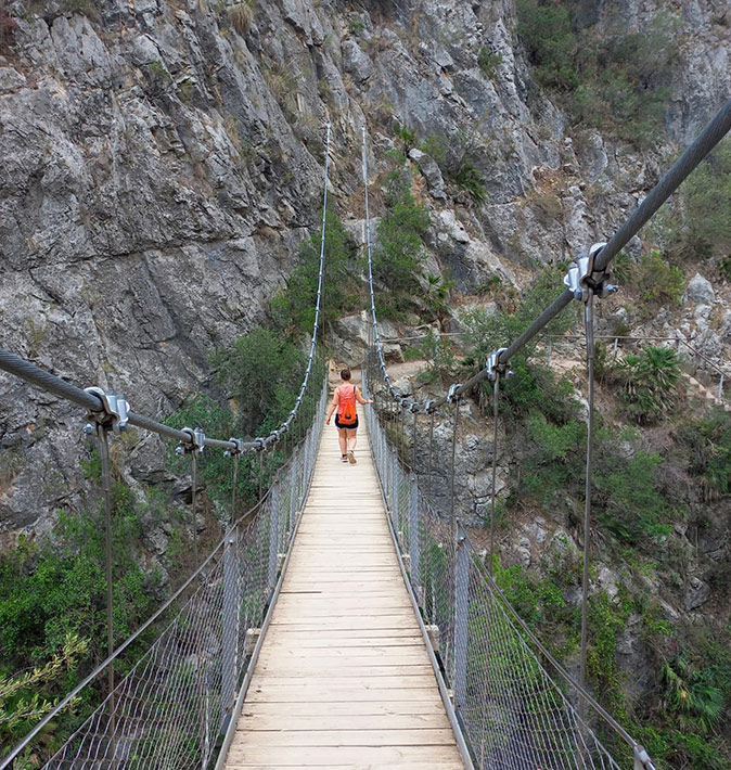A woman crossing a suspension bridge during a hike surrounded by mountains and some greenery
