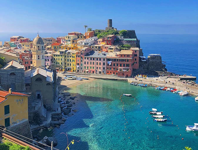 a bay with several boats in the foreground, a small beach, colorful houses and a castle on a hill in the background