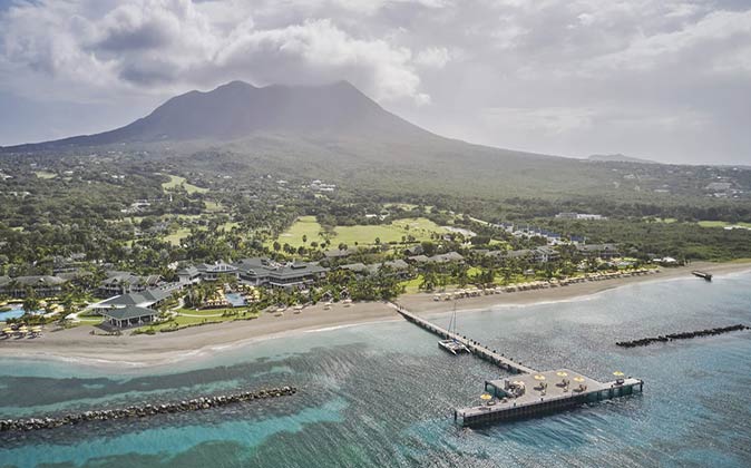 Four Seasons Resort Nevis, one of the best luxury resorts in the Caribbean aerial view of a Caribbean island with the beach and a luxury resort in the foreground and green hills and a mountain covered in clouds in the background