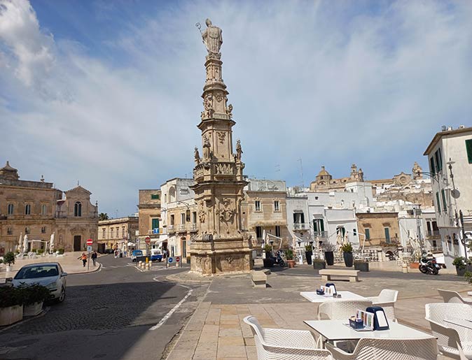 visit places outside of high season to stay more off the beaten path a tall religious statue in the center of a large town square, with white tables and chairs in the foreground and whitewashed houses in the background