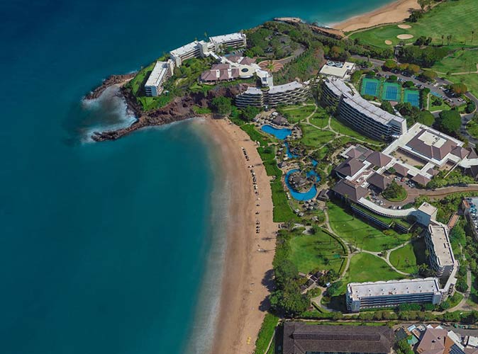 an aerial view of Ka'anapali Beach, a long sandy beach with a dark blue sea and a resort with swimming pools in the background