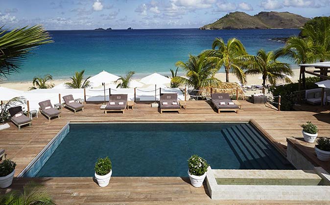 a swimming pool surrounded by a wooden deck and luxury sun loungers, with a white sandy beach and azure blue sea in the background
