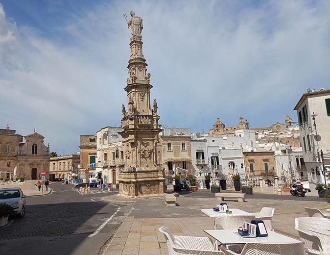 white chairs and tables on a square with a tall statue and brown and white buildings