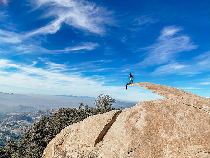 a woman sitting on a thin piece of rock overlooking the valley