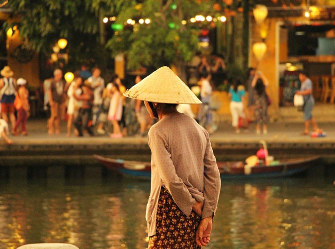 woman wearing a traditional Vietnamese hat, looking out over a river with a lot of people on the other side