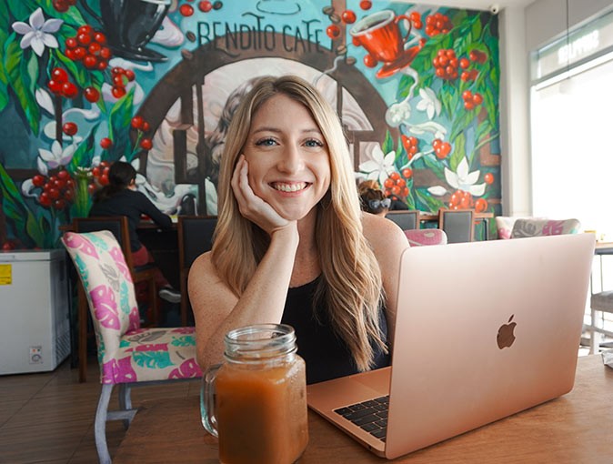 A woman sitting in a cafe smiling at the camera with a laptop and iced coffee on the table